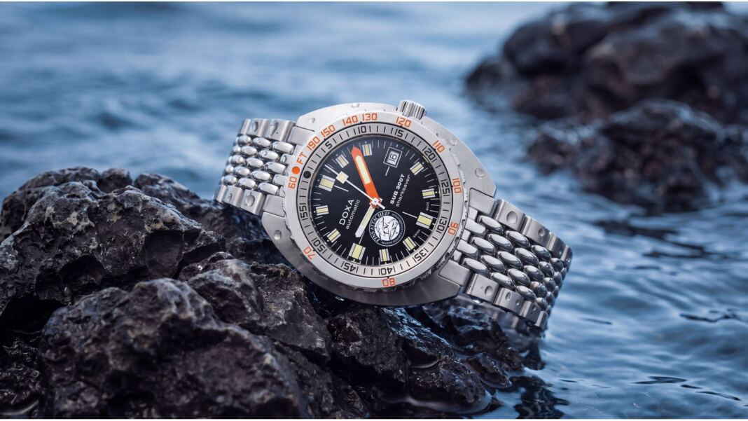 doxa-sub-300t-sharksaver-fins-attached-hero-1600x900-1 DOXA SUB 300T Sharksaver Fins Attached edition on a rocky shoreline, steel beads-of-rice bracelet