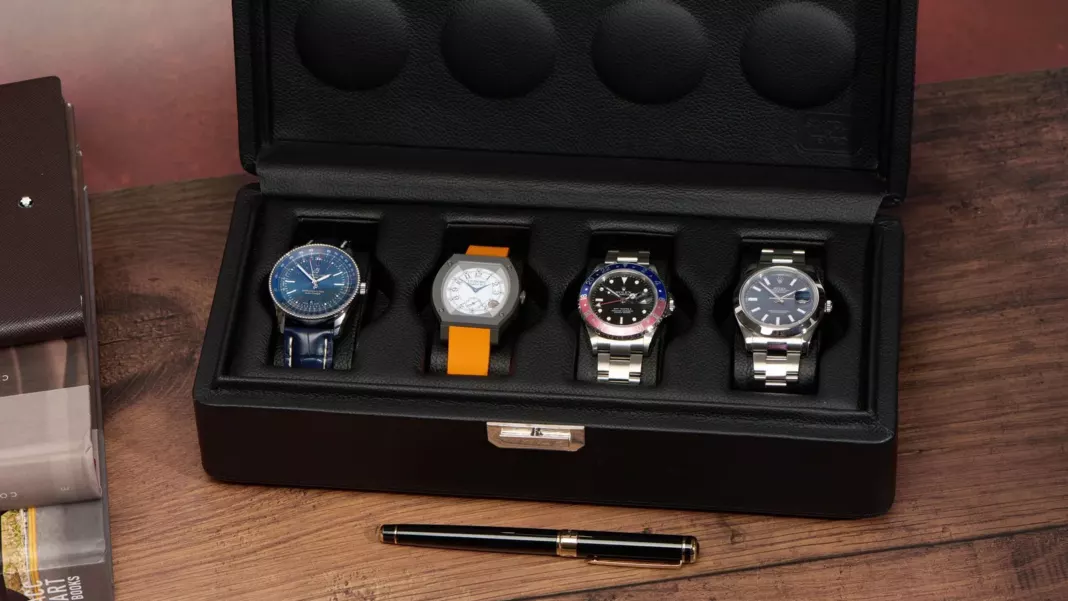 Four watches in an open black leather watch box on a wooden desk